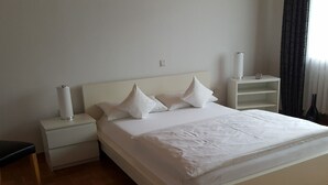 A bedroom with a large bed, white bedding, a nightstand, a bedside lamp, and a built-in shelf.