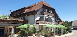 Ein zweistöckiges Gebäude mit einem Schild, auf dem 'Pension Linde' steht. Es gibt Tische und Stühle im Freien sowie Sonnenschirme.