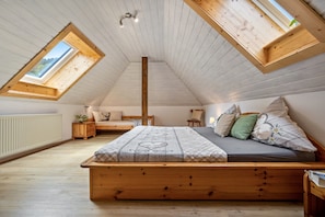 A loft bedroom with a sloped ceiling, wooden beams, a central skylight, a bed with a patterned bedspread, and a view of the outdoors.