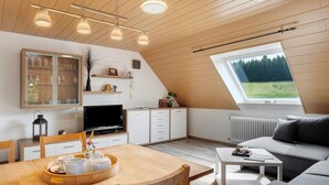 A modern loft-style room with a dining area, a sofa, a TV, and a large window with a view of trees. - Apartment 'Ringwald' with Mountain View, Shared Garden and Wi-Fi (Biederbach)