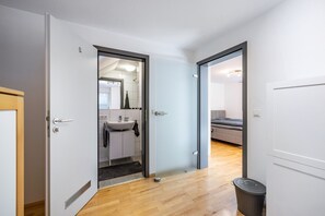 A modern interior with a wooden floor, a white door leading to a bathroom, and a visible bed in the adjacent room.