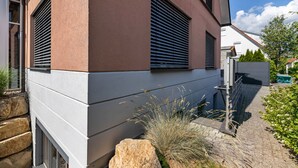 A modern building with a stone base, large windows with horizontal blinds, and a paved pathway with plants.