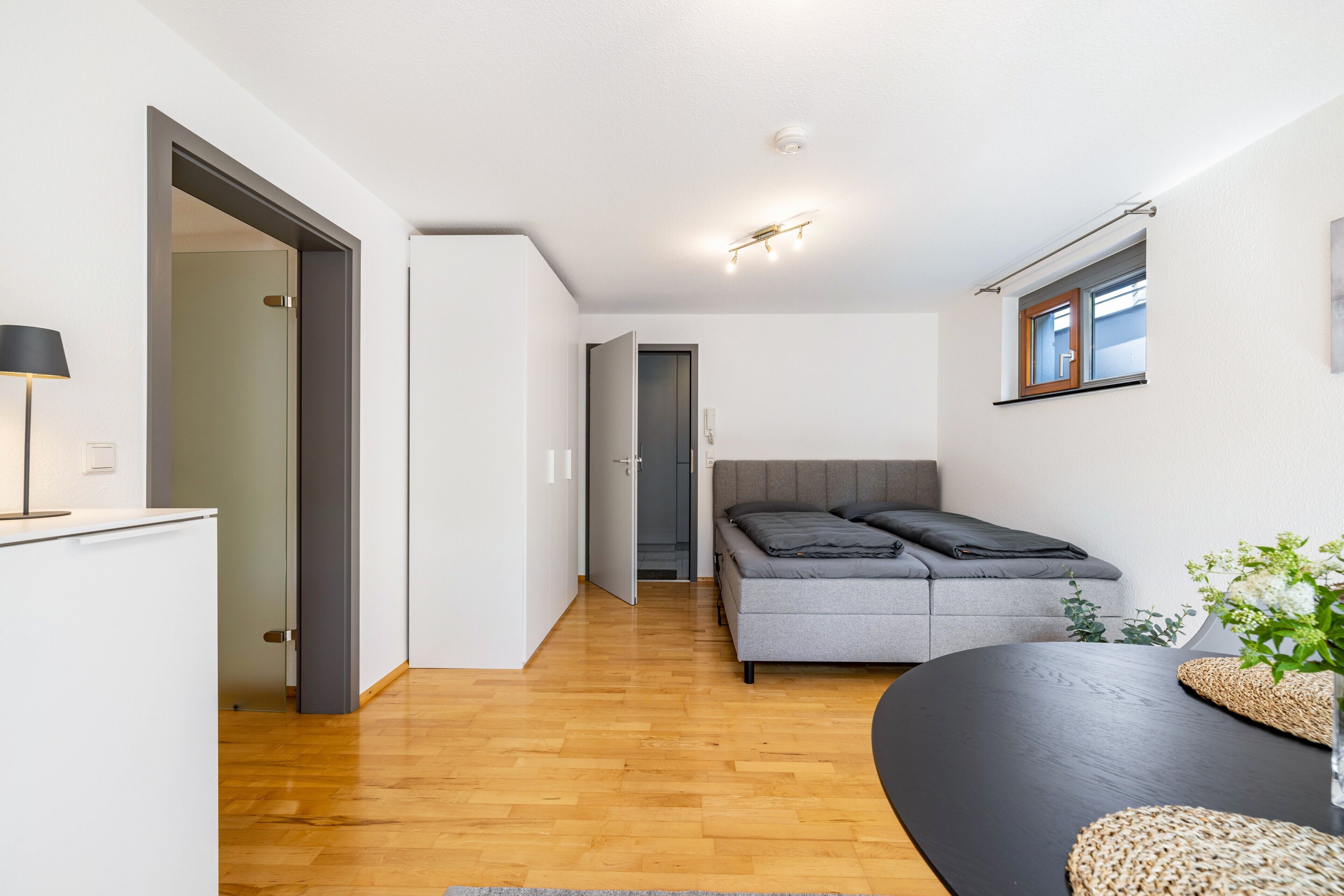 A modern, compact room with a wooden floor, a grey sofa, a small table, and a window with a view.