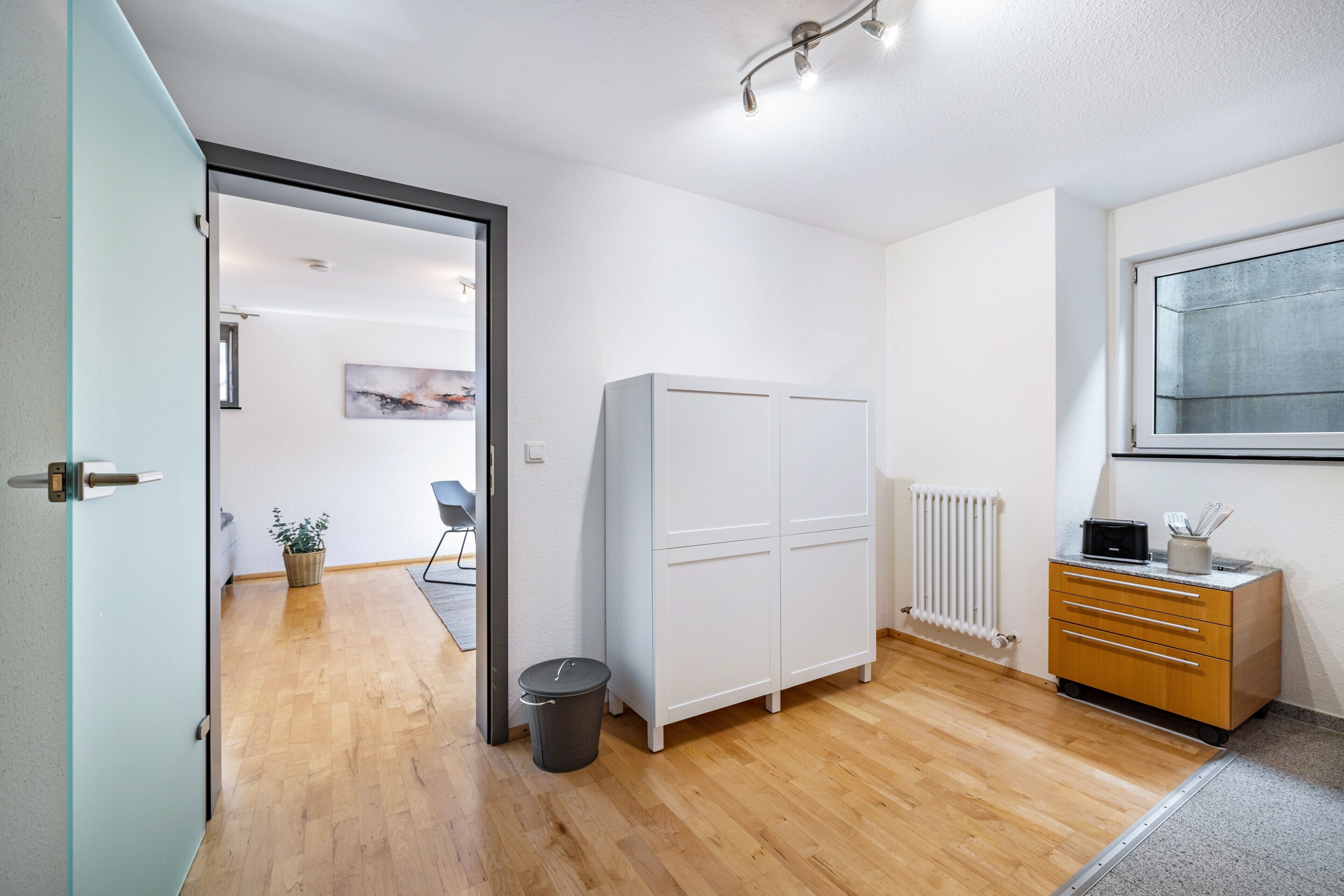 Ein Zimmer mit Holzboden, einem weißen Kleiderschrank, einem Heizkörper, einer kleinen Kommode und einem Fenster.