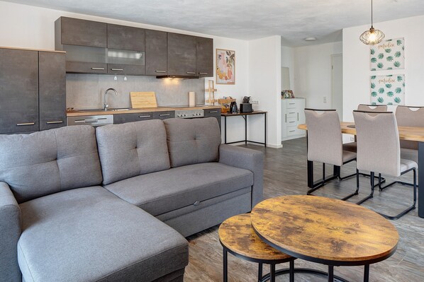 Une cuisine moderne avec des meubles de couleur sombre, un canapé gris et un coin repas comprenant une table et des chaises en bois.