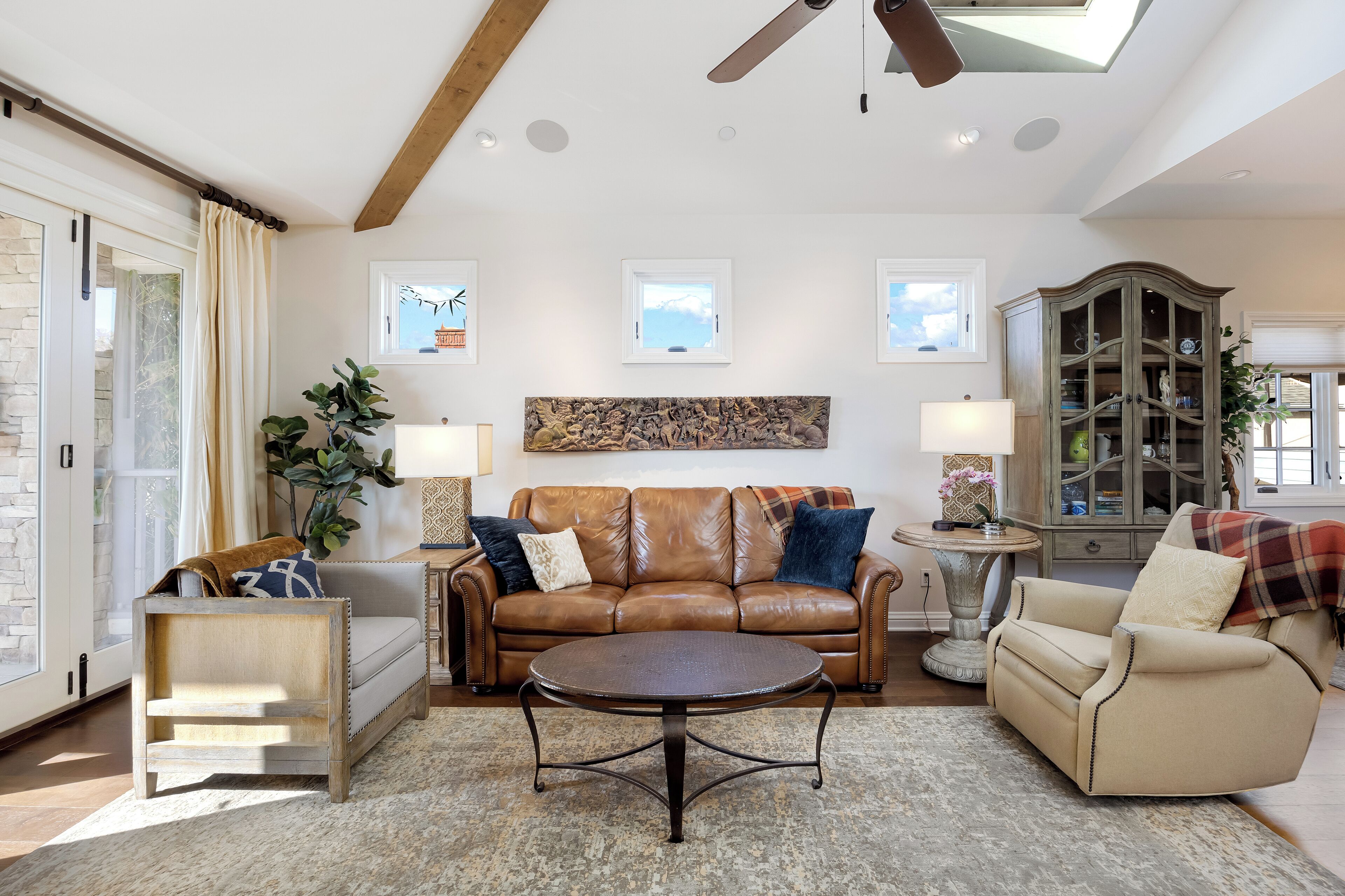 A spacious living room with a brown leather sofa, a round coffee table, and a beige armchair. There is a wooden cabinet with glass doors and a ceiling fan.