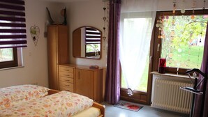 A bedroom with a bed, wooden wardrobe, mirror, window with curtains, radiator, and a view of greenery outside. - Apartment 'Bienenherz' with Private Terrace, Shared Garden, and Wi-Fi (Engelsbrand)