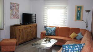 A living room with a sofa, ottoman, coffee table, TV, wooden cabinet, and artwork on the walls. - Apartment 'Bienenherz' with Private Terrace, Shared Garden, and Wi-Fi (Engelsbrand)