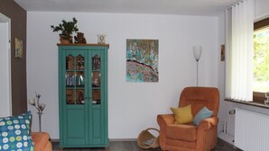 A living room with a teal cabinet, an orange armchair, a blue patterned pillow, and a painting on the wall. - Apartment 'Bienenherz' with Private Terrace, Shared Garden, and Wi-Fi (Engelsbrand)