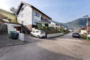 Une rue résidentielle bordée de maisons, avec une voiture blanche garée sur le côté et une autre voiture visible au loin.