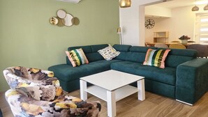 Una sala de estar con un sofá verde, una mesa de centro blanca y un sillón estampado. Se ve un comedor al fondo.