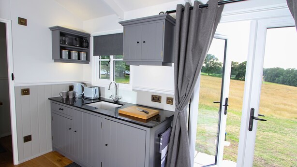 Une cuisine de taille réduite avec des meubles gris, un plan de travail en bois et une fenêtre agrémentée de rideaux.
