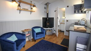 Un espace de vie compact comprenant un coin cuisine, deux fauteuils bleus, une cheminée et une petite table à manger.