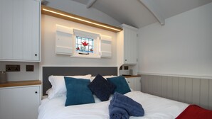 Un lit bien fait, avec du linge de lit blanc, des coussins bleus et turquoise, et un plaid rouge. On y trouve une tête de lit en bois, une table de chevet blanche avec une lampe, et une fenêtre ornée de vitraux au-dessus du lit.