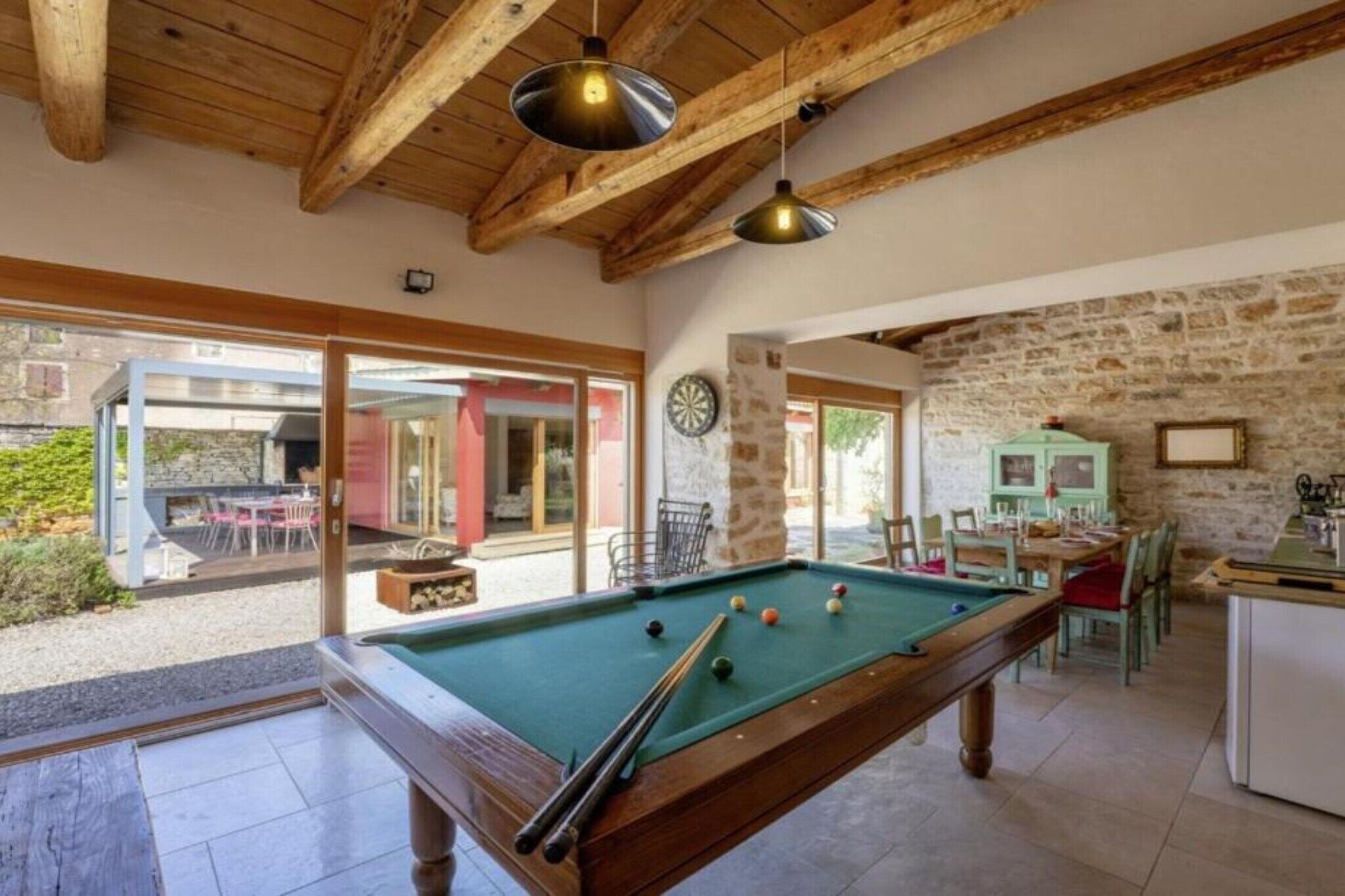 Una mesa de billar en una sala con paredes de piedra y vigas de madera, un comedor con sillas, y vistas al exterior.