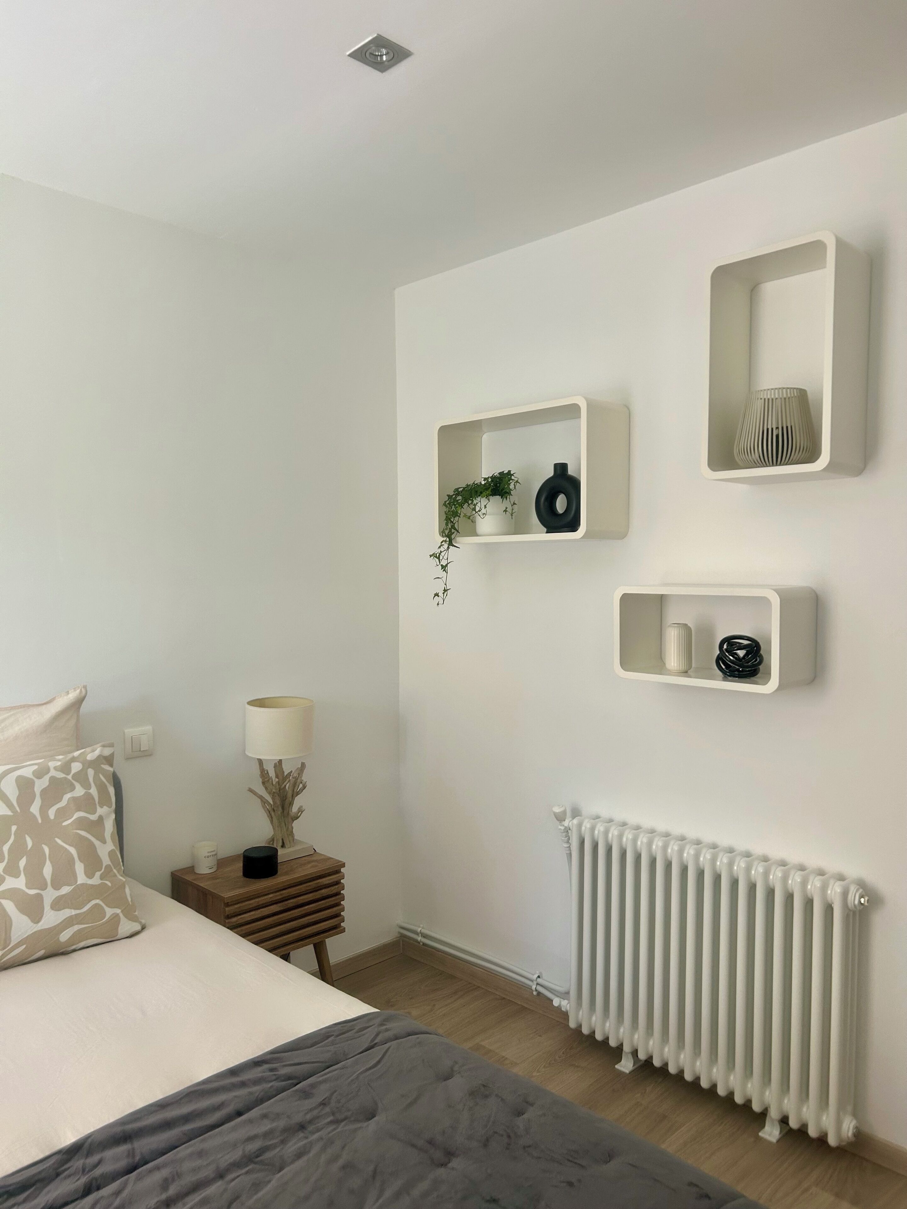 A bedroom with a bed, bedside table, lamp, radiator, and wall-mounted shelves with decorative items.