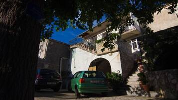 Une rue étroite où est garée une voiture verte sur le bord, un bâtiment en pierre doté d’un balcon et un arbre qui offre de l’ombre.