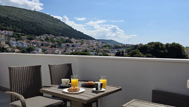 Een balkon met rieten stoelen en een tafel, gedekt met ontbijtspullen en drankjes, met uitzicht op een heuvelstad.