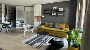 A modern living room with a sofa, a coffee table, and a black cat. There is a kitchen in the background and a plant on the right. - Modern & Charming - Free Private Parking - One Bedroom Apartment (Zagreb)