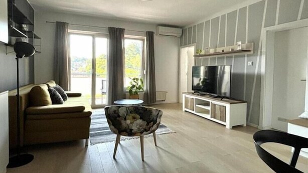 A modern living room with a sofa, a floral chair, a TV, and a balcony with curtains. - Modern & Charming - Free Private Parking - One Bedroom Apartment (Zagreb)