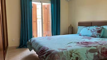 Un dormitorio con cama, cortinas azul verdosas, una ventana con contraventanas de madera y un aire acondicionado fijado a la pared.