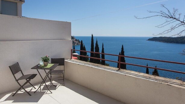 Un balcon avec une table et des chaises, offrant une vue sur la côte, avec des cyprès et la mer.
