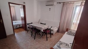 Una habitación pequeña y limpia con una mesa de comedor, sillas, una cama y una ventana con cortinas.