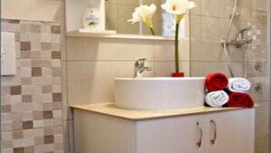 A bathroom with a white sink, a mirror, and a shower. There are towels on the sink and a red vase with flowers. - Sea View Apartments Lendic Podgora - Comfort Studio Apartment with  Balcony and Sea View (Podgora)