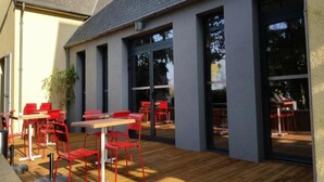 Een buitenruimte met rode stoelen en tafels, grote glazen deuren en een houten terras.