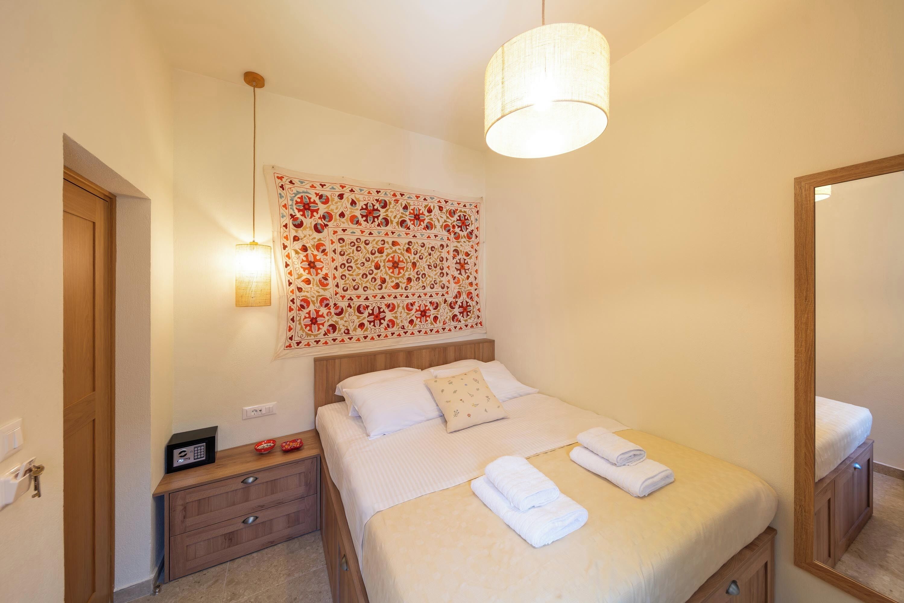 A neatly made bed with white linens and a folded towel, a bedside table with a lamp, a wall-mounted artwork, and a mirror.