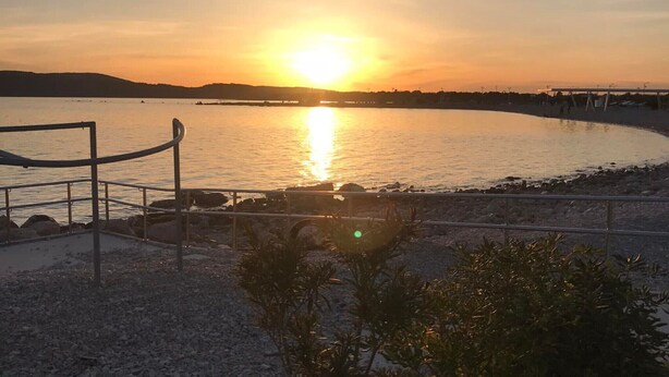 A sunset over a calm body of water with a beach and a fence in the foreground. - Apartment Belu - Two Bedroom Apartment With Sea View (Brodarica)