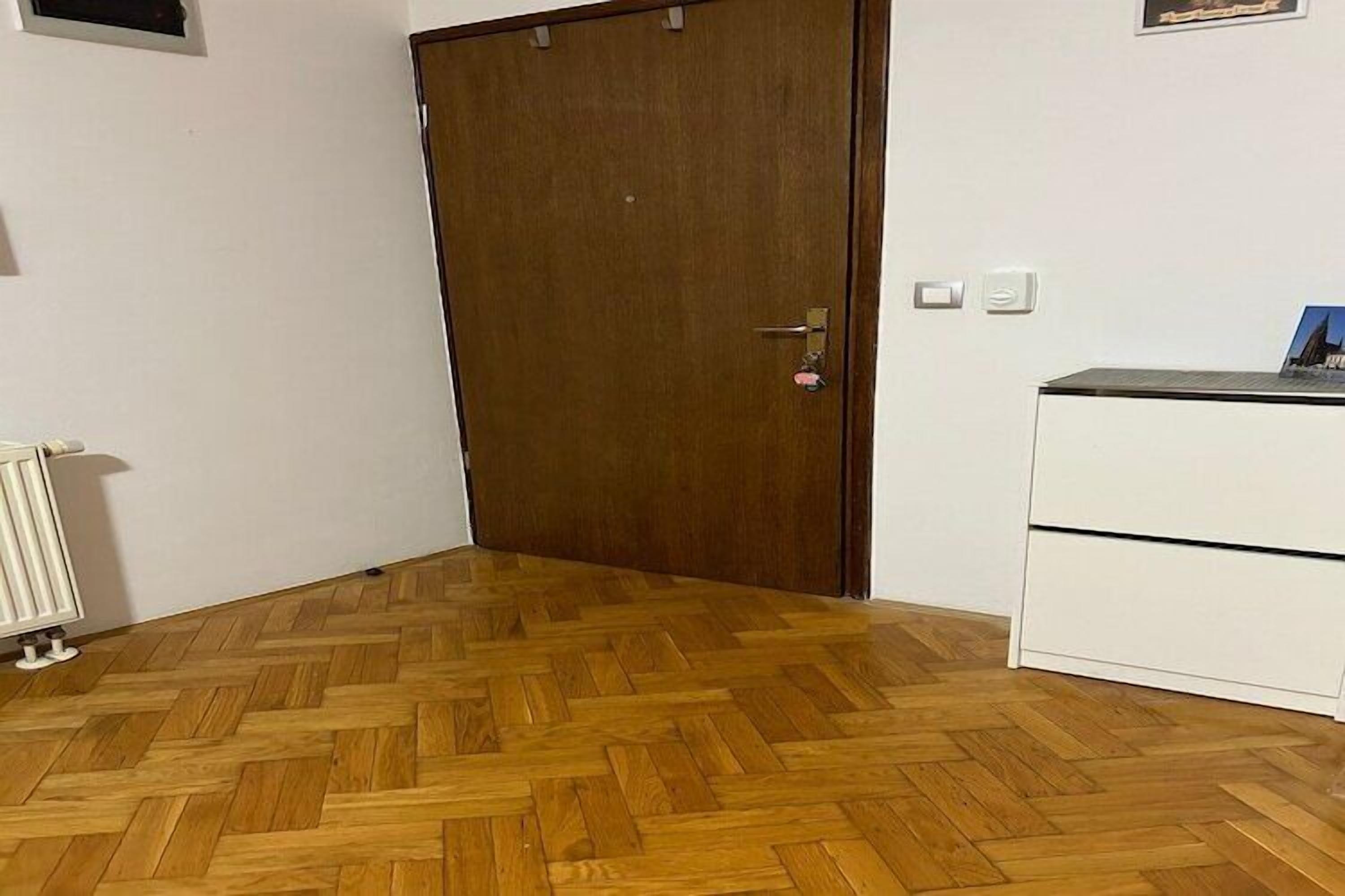 Una stanza con pavimento in parquet di legno, una porta in legno chiusa, un termosifone bianco e un armadietto bianco con cassetti.