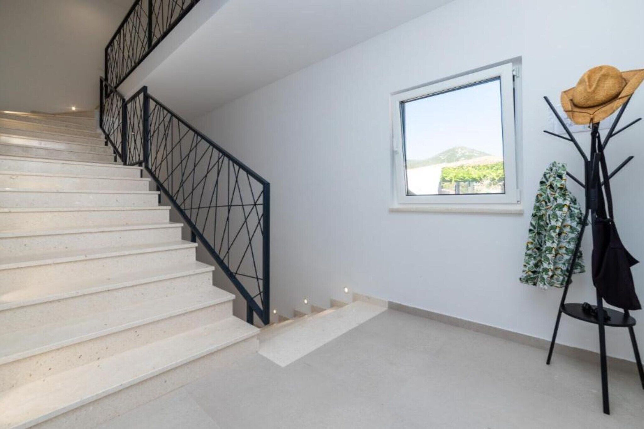 Eine Treppe mit geometrischer Geländerkonstruktion, ein Fenster mit Aussicht und ein Kleiderständer mit Hüten und einer Jacke.