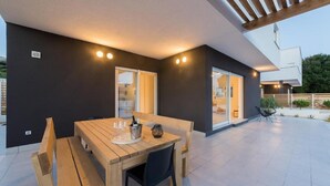 An outdoor patio with a wooden table and chairs, a pergola, and a modern building with large windows. - Luxury Villas Bellavista - Four Bedroom Villa with Pool (Pag)