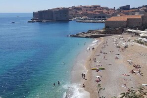 A beach with people sunbathing and swimming, a historic building in the background, and clear blue waters.