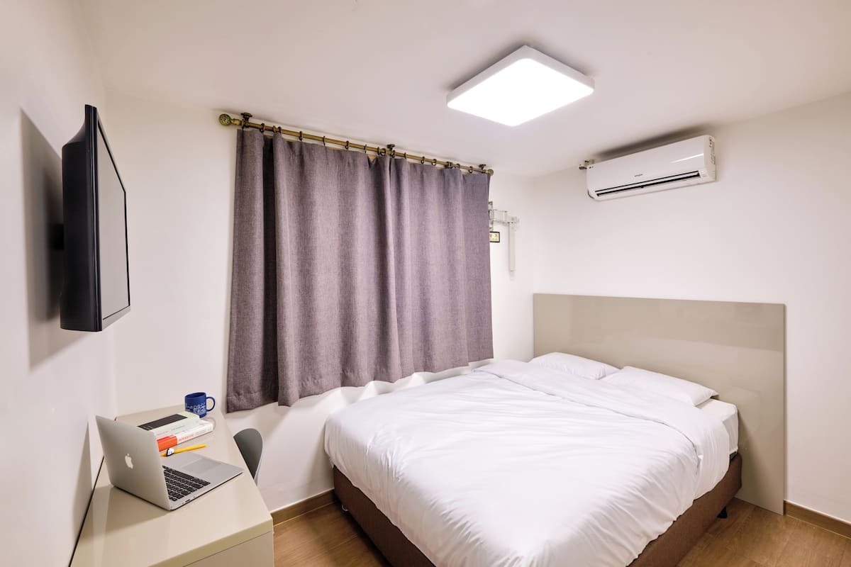 A hotel room with a bed, a desk with a laptop, a flat-screen TV, and an air conditioner.