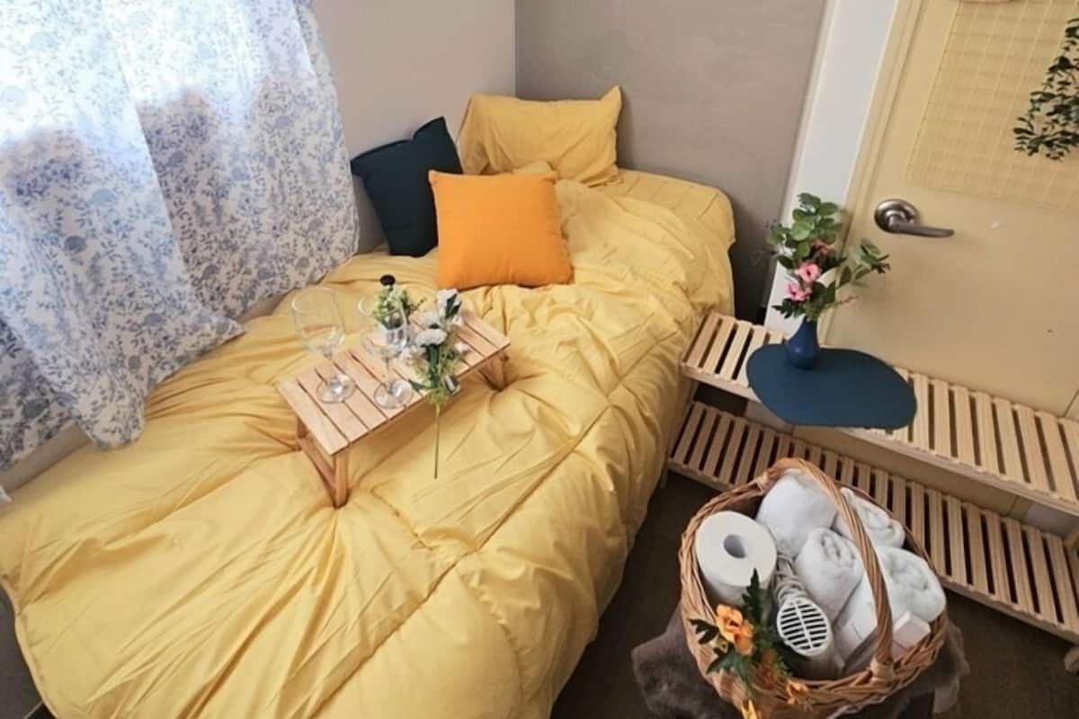 A neatly made bed with a yellow duvet, a bedside table with a vase and a basket, and a wooden bench with a towel.