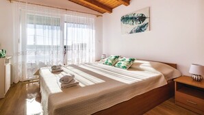 Um quarto com teto de madeira, cama com colcha branca e almofadas verdes, mesa de cabeceira com abajur, e janela com cortinas finas.