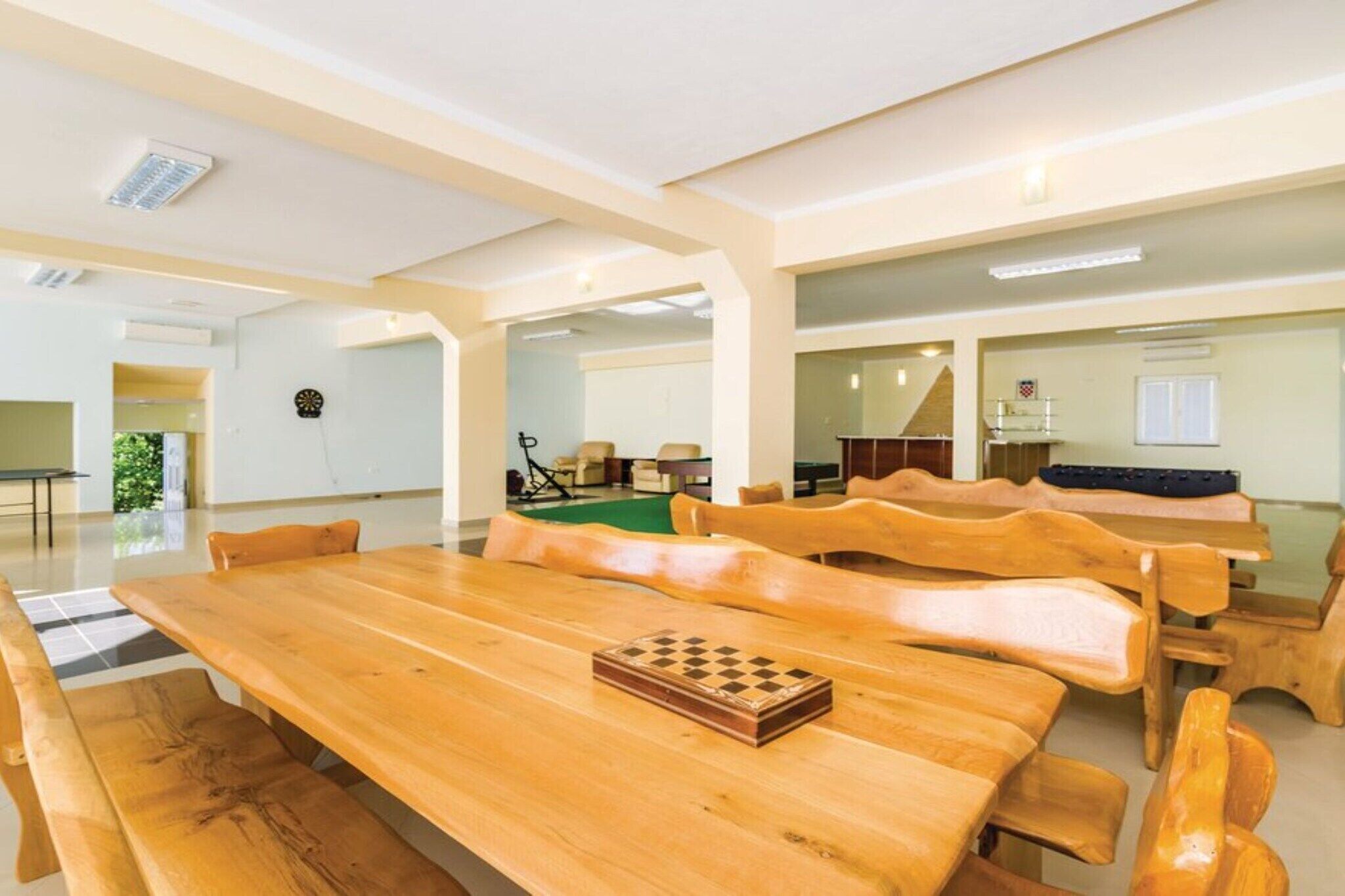 Um quarto espaçoso com uma longa mesa de madeira e bancos, um alvo de dardos e um tabuleiro de xadrez.