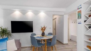 Una sala moderna con una televisión de pantalla plana montada en la pared, una mesa de comedor redonda con sillas, un sofá con cojines y un estante blanco con diversos objetos.