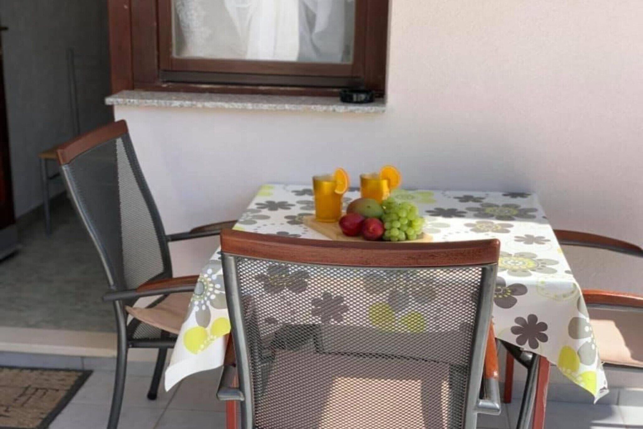 Une table à manger recouverte d’une nappe à motifs floraux, une chaise et un panier de fruits contenant des oranges, des raisins et des pommes.