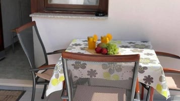 Une table à manger recouverte d’une nappe à motifs floraux, une chaise et un panier de fruits contenant des oranges, des raisins et des pommes.