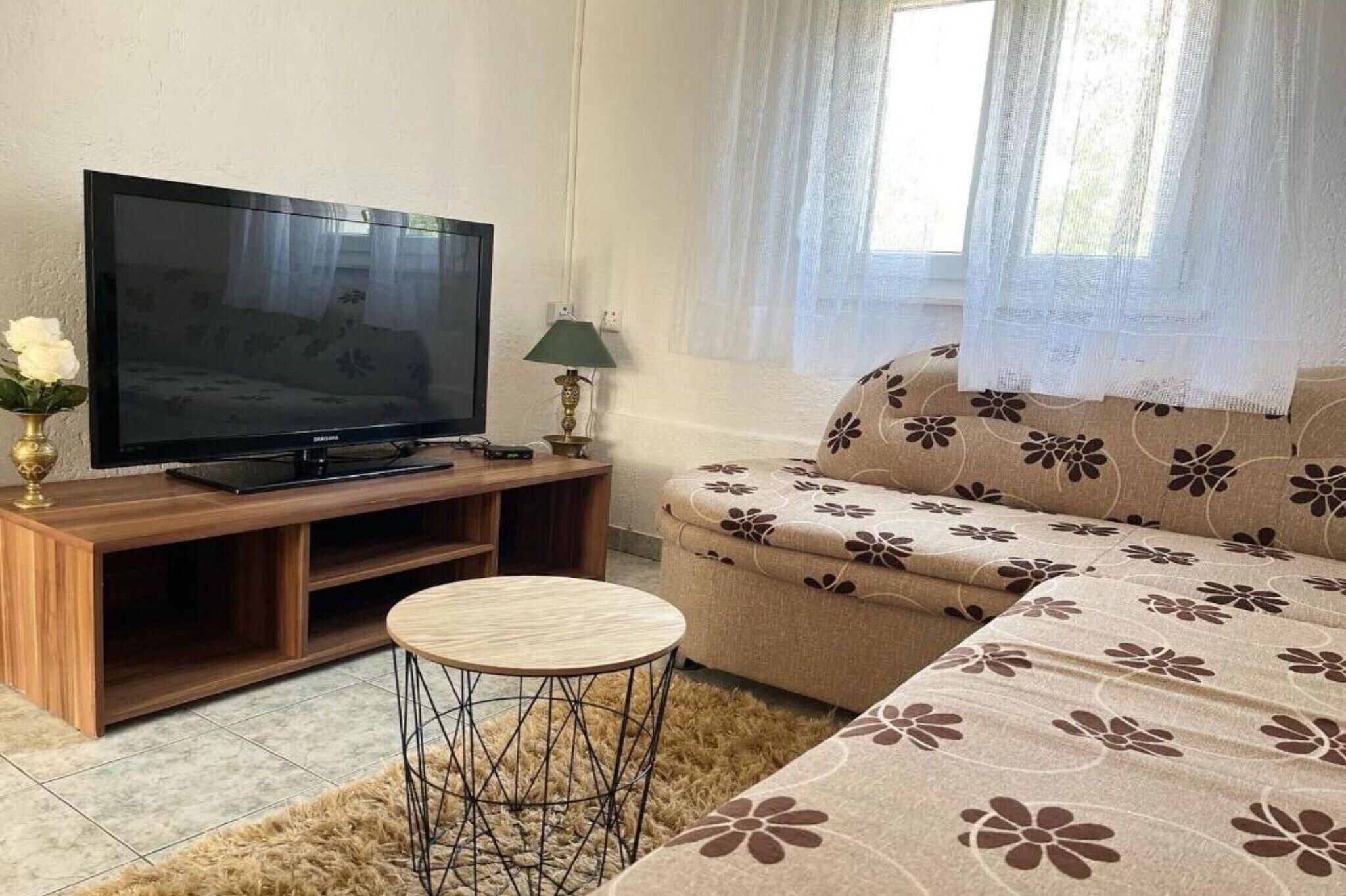 Ein Wohnzimmer mit Fernseher auf einem Holzboden, einem runden Couchtisch, einer Couch mit Blumenmuster und einem Fenster mit durchscheinenden Vorhängen.