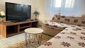 A living room with a television on a wooden stand, a round coffee table, a sofa with a floral pattern, and a window with sheer curtains. - One Bedroom Apartment with Balcony (3) (Biograd na Moru)