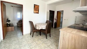 A dining area with a round table and chairs, a kitchen with a stove and sink, and a hallway leading to a bedroom. - One Bedroom Apartment with Balcony (3) (Biograd na Moru)