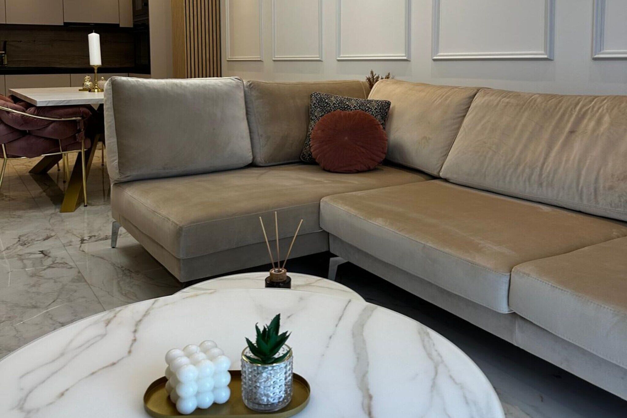 A modern living room with a beige sectional sofa, a round marble coffee table with decorative items, and a dining area with a white table and gold chairs.