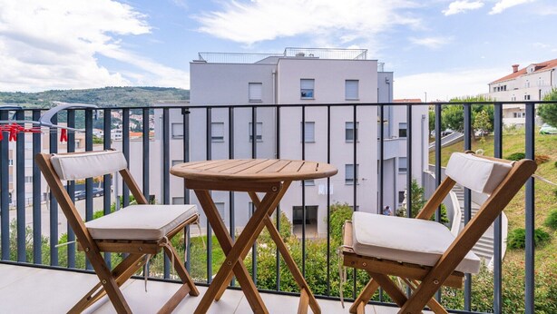 Un balcon avec deux chaises en bois et une petite table, donnant sur un quartier résidentiel avec des immeubles et de la verdure.