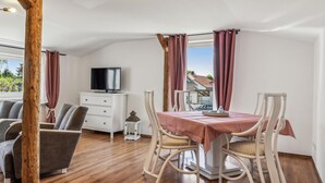 A dining area with a table set for two, two chairs, a television on a white cabinet, and a window with curtains.