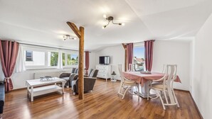 A spacious room with a wooden floor, a dining table with chairs, a sofa, a TV, and a window with curtains.
