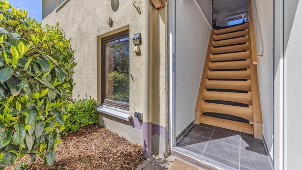 A house with a wooden staircase leading to a glass door, a window, and a small garden area with plants.
